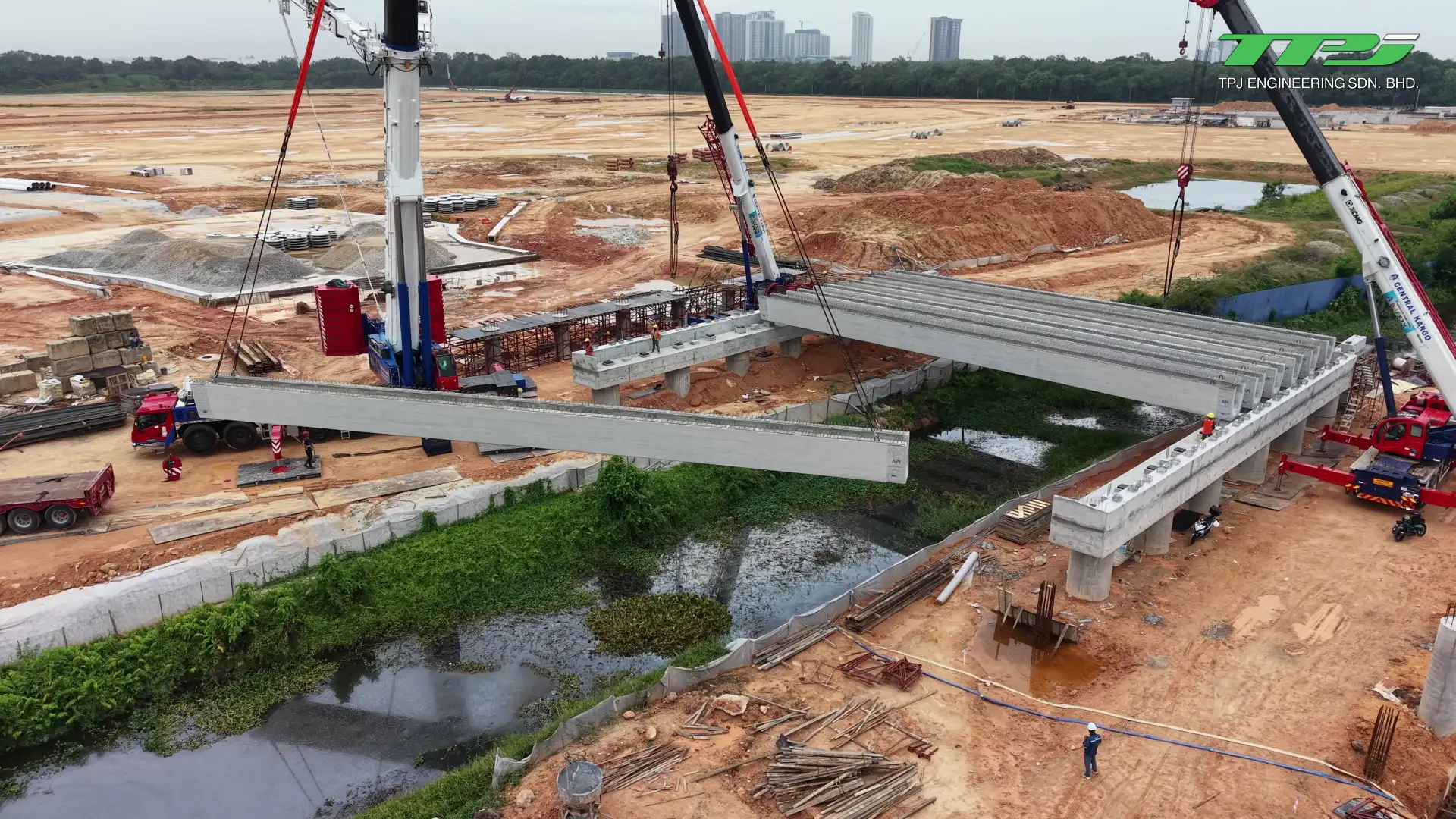 Cranes lifting a precast U-beam across Sungai Binjai beside installed beams at the Batu Kawan bridge site.