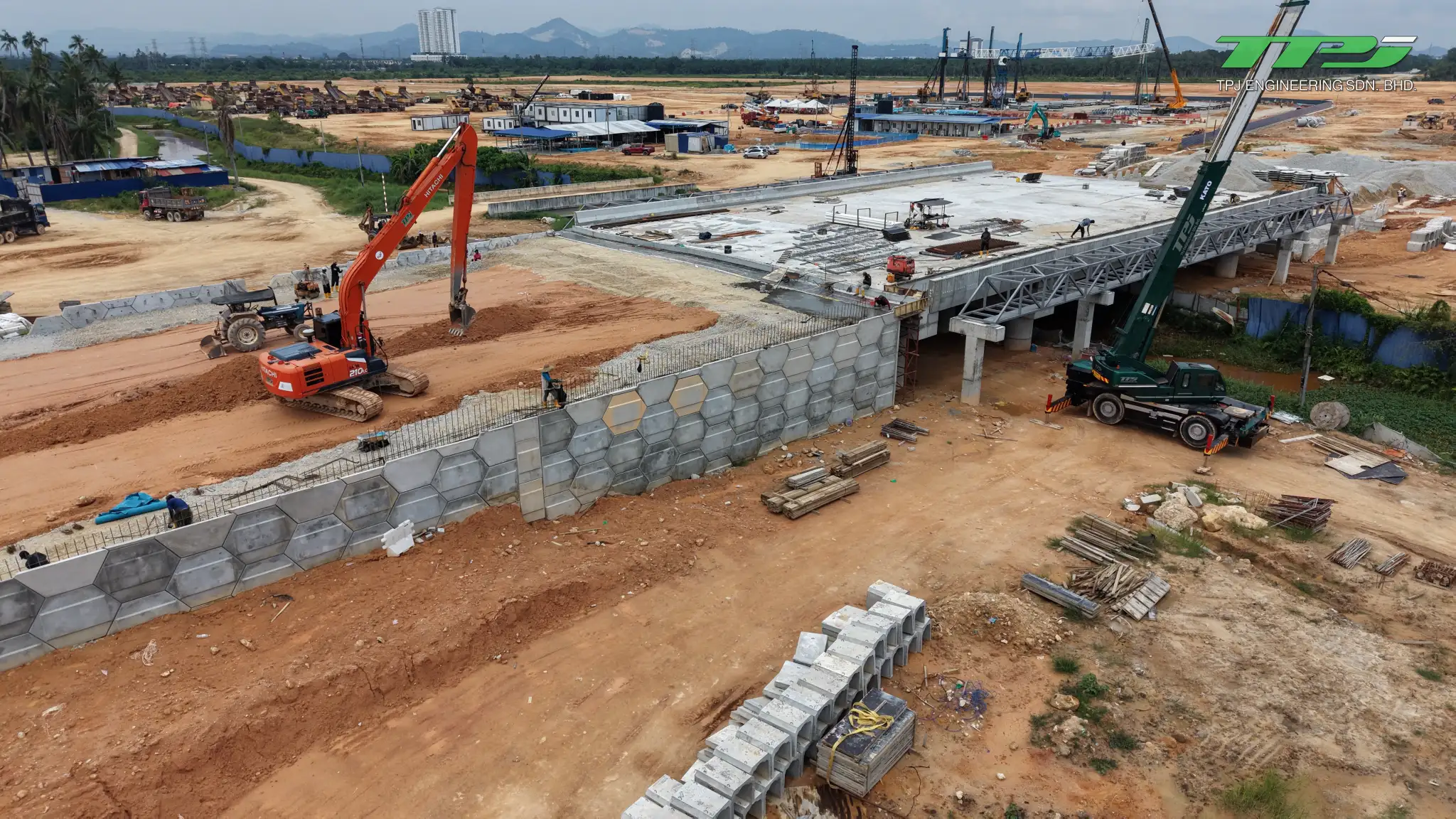 Hexagonal retaining wall panels beside the Batu Kawan bridge deck, with an excavator and crane.